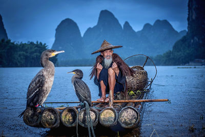 Man with birds sitting on wooden raft in lake
