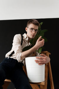 Portrait of young man embracing plant while sitting on ladder against wall