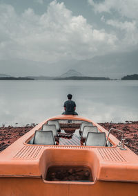 Rear view of man sitting on boat by lake against cloudy sky