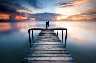 Couple standing on pier over sea against sky during sunset