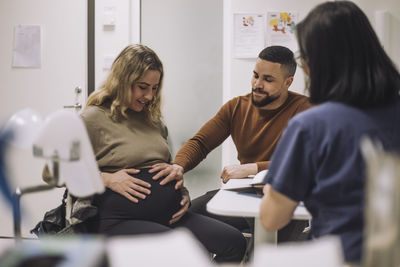 Smiling man touching stomach of pregnant woman while discussing with gynecologist in medical clinic