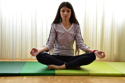 Woman meditating while sitting on exercise mat against curtains at home