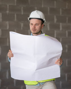 Portrait of young man holding hardhat while standing against wall