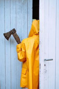 A person in a yellow raincoat and a rusty ax in his hands is hiding behind the ajar door of the barn