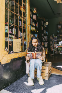 Girl reading book while sitting on wooden crate near door in bookstore