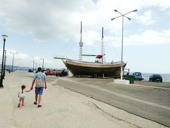 Rear view of father and daughter walking on street by harbor against cloudy sky