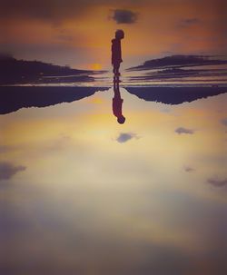 Symmetry view of boy standing on shore at beach against sky during sunset