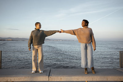 Friend stretching hands while standing at distant on bench against sea