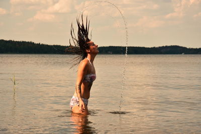 Woman tossing hair while standing in lake against sky