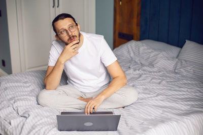 Man in a white t-shirt, a freelancer with glasses, sits on the bed at home with a laptop and works 