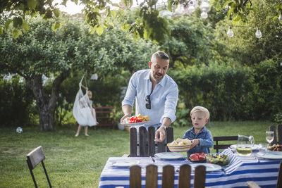Mature father directing son with food plate while pointing at table in yard
