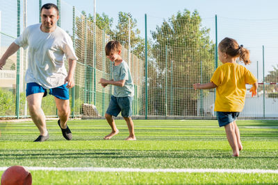 Man with children playing football on green sports field.