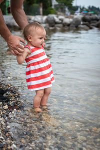 Portrait of baby girl in striped dress standing in water of the lake being held by father hands