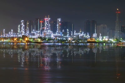 Illuminated modern buildings by river against sky at night