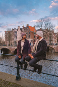 Full length of couple sitting on railing by canal against buildings