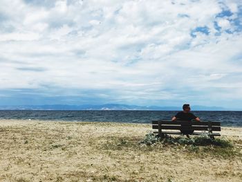 Man sitting on bench at beach against sky