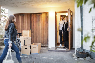 Parents looking at daughter walking by cardboard boxes and bicycle outside new house