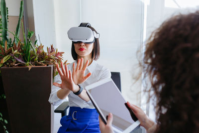 Sitting asian woman wearing viewer exploring virtual reality with her colleague working with tablet