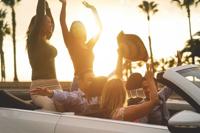 Happy friends with arms raised traveling in convertible against sky during sunset