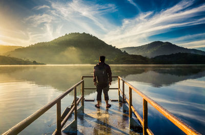 Rear view of man standing on railing by lake against sky