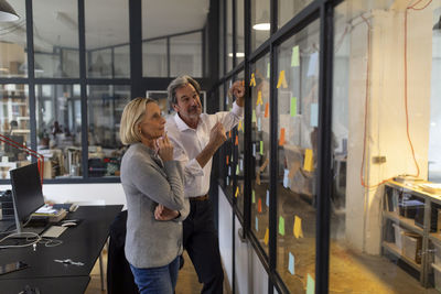Colleagues looking at sticky notes at glass pane in office
