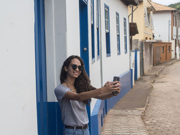 Smiling young woman taking selfie while standing by wall