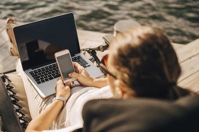 High angle view of woman with laptop text messaging on smart phone while sitting at jetty