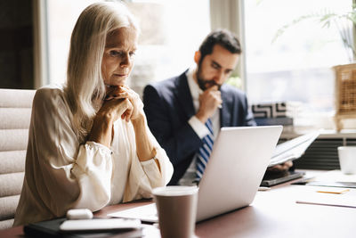 Mature businesswoman staring at laptop by businessman at desk in office