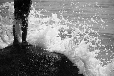 Low section of man standing on rock while water splashing at sea shore