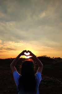 Rear view of woman making heart shape while standing against sky during sunset