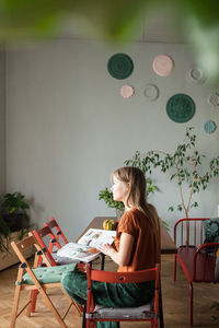 Woman sits at table with magazine, looks at window in living room. person