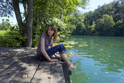 Young woman sitting by lake against trees