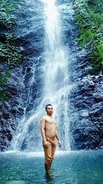 Portrait of shirtless man standing against waterfall in forest