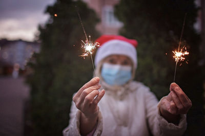 Portrait of senior woman wearing mask holding sparklers against trees