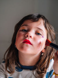 Kid with applicator making up face with assorted cosmetic products in house looking at camera