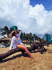 Full length of young woman sitting on fallen tree at beach