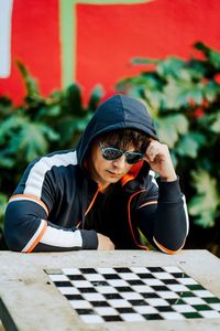 Man wearing a  hoodie and sunglasses looking down at a chessboard with a colorful wall as background
