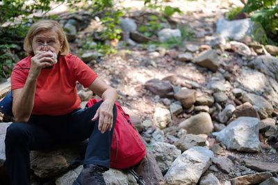 Senior woman sitting on the rock in nature and eating sandwich