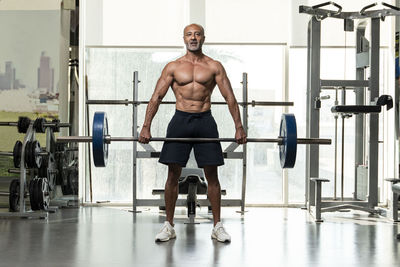 Full length of shirtless man holding barbell standing in gym