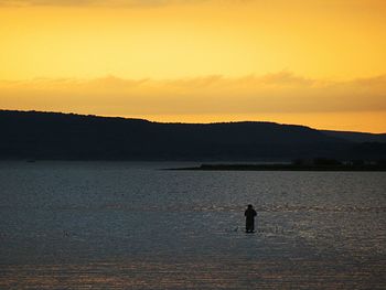 Silhouette man fishing in lake against orange sky at sunset