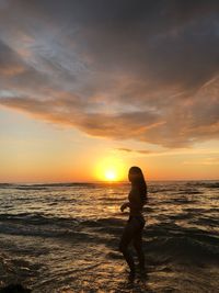 Side view of woman in bikini standing at beach against sky during sunset
