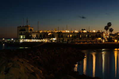 Illuminated buildings by sea against sky at night