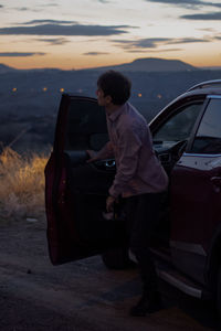 Side view of man standing on car at sunset