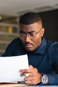 Serious black adult male in formal clothes and eyeglasses sitting at table while taking notes in notebook from papers near netbook in bright workplace