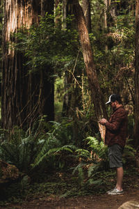 Full length of young man standing by tree in forest operating drone remote control looking up