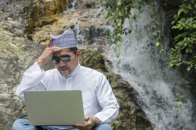 Mature man wearing sunglasses while using laptop against waterfall