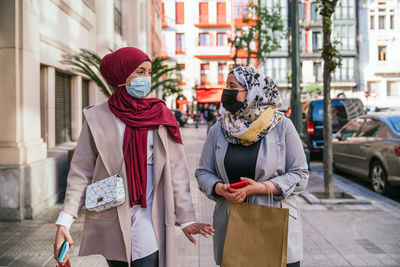 Muslim female friends in masks and with paper bags walking in city after shopping looking at each other