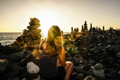 Rear view of woman sitting by stone stacks at sea shore against sky during sunset