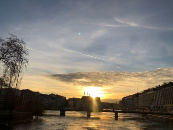 Silhouette bridge over river by buildings against sky during sunset