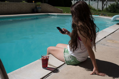 Young woman using mobile phone while sitting by swimming pool during summer